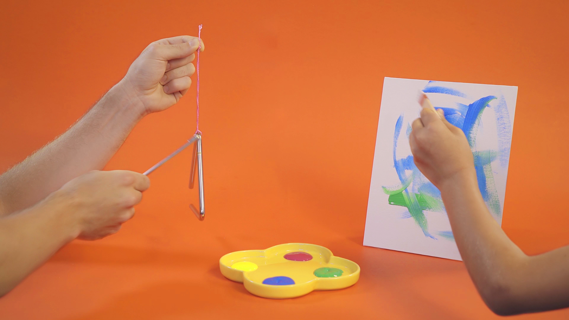 À gauche, il y a deux mains qui jouent du triangle, au centre on retrouve un plat avec de la peinture, et à droite une main qui peint un tableau
