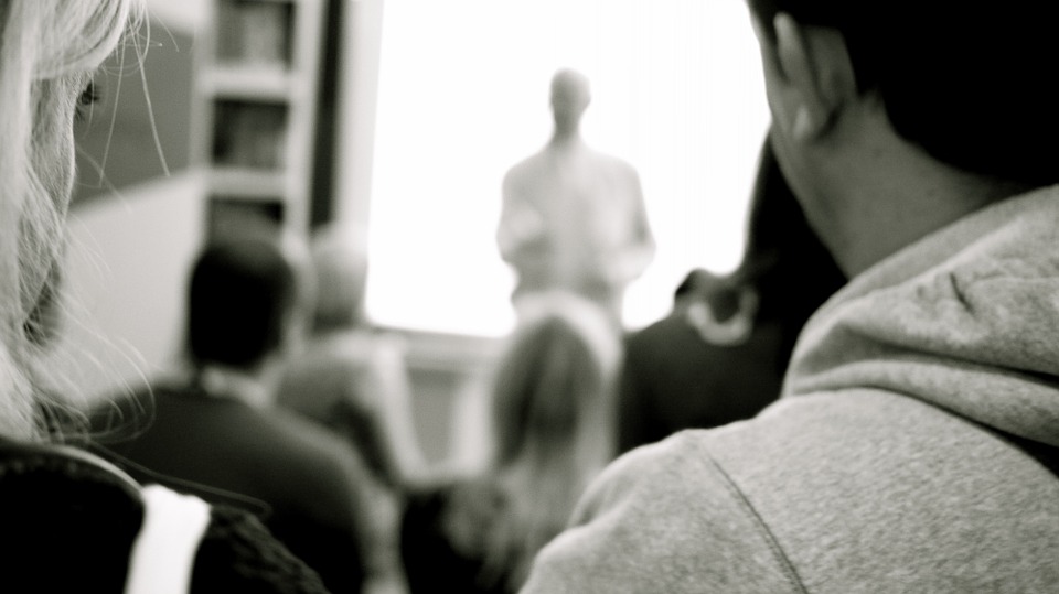 Photo en noir et blanc, deux étudiants sont de dos. Ils regardent le professeur.